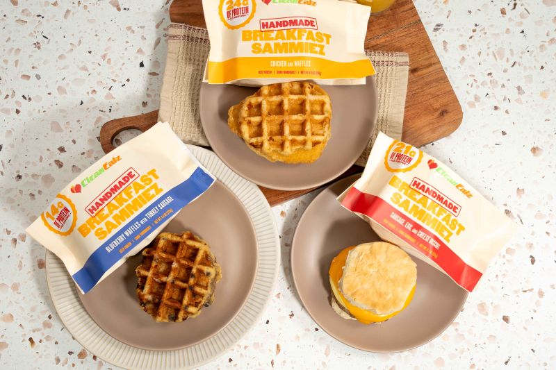 Top-down close-up of three Breakfast Sammiez on plates with matching Clean Eatz Handmade Breakfast Sammiez packages: Chicken & Waffles, Blueberry Waffles with Turkey Sausage, and Sausage, Egg & Cheese Biscuit.