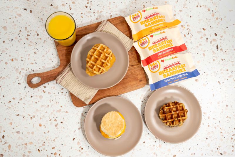 Top-down view of three plated Breakfast Sammiez (waffle and biscuit sandwiches) with a glass of orange juice and three Clean Eatz Handmade Breakfast Sammiez packages on a countertop.