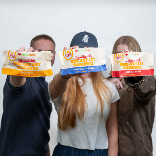 Three people holding Clean Eatz Handmade Breakfast Sammiez packages toward the camera: Chicken & Waffles, Blueberry Waffles with Turkey Sausage, and Sausage, Egg & Cheese Biscuit.