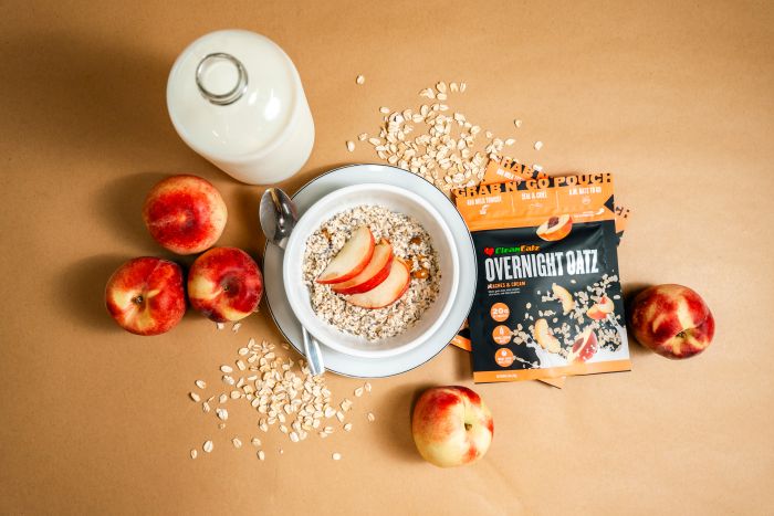 Top-down shot of a Peaches & Cream Overnight Oatz setup with a bowl of oats topped with sliced peaches, a milk bottle, fresh peaches, scattered oats, and the Overnight Oatz pouch on a tan background.
