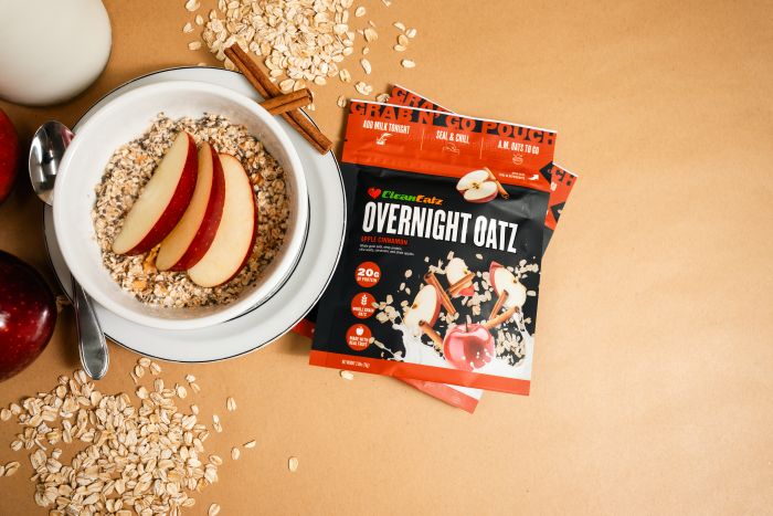 Top-down view of a bowl of overnight oats topped with sliced apples next to a Clean Eatz Overnight Oatz pouch, with oats scattered on a tan surface.