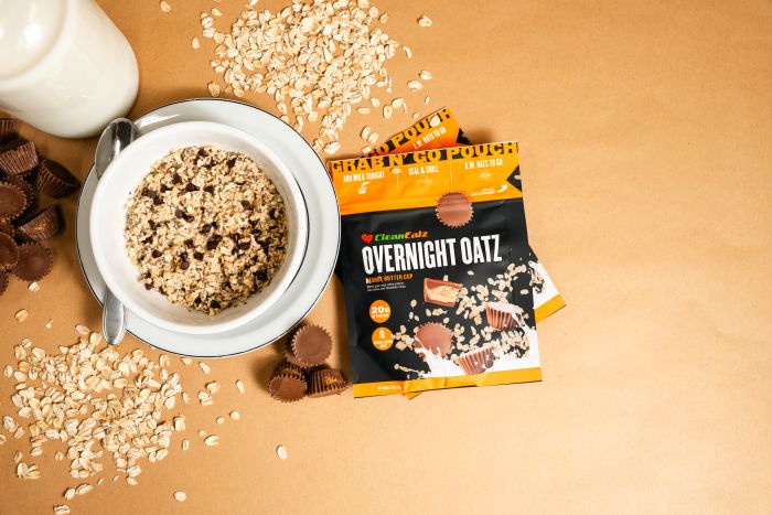 Top-down shot of a Peanut Butter Cup Overnight Oatz setup with a bowl of oats, mini peanut butter cups, scattered oats, and the Overnight Oatz pouch on a tan background.