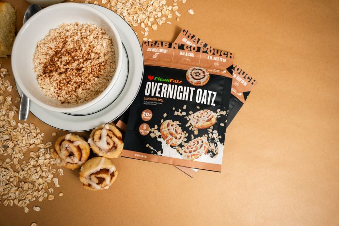 Top-down view of a bowl of overnight oats with cinnamon-roll bites beside a Clean Eatz Overnight Oatz Cinnamon Roll pouch, with oats scattered on a tan surface.