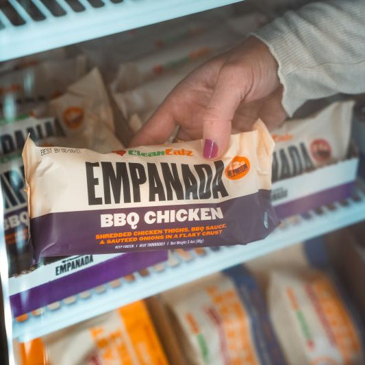 Hand holding a Clean Eatz Empanada BBQ Chicken package in front of a freezer shelf stocked with empanadas.