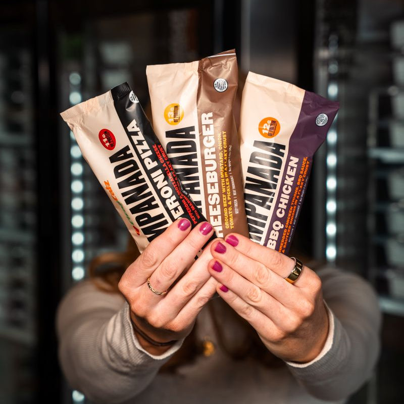Person holding three Clean Eatz Empanada packages toward the camera—Pepperoni Pizza, Cheeseburger, and BBQ Chicken—with freezers in the background.