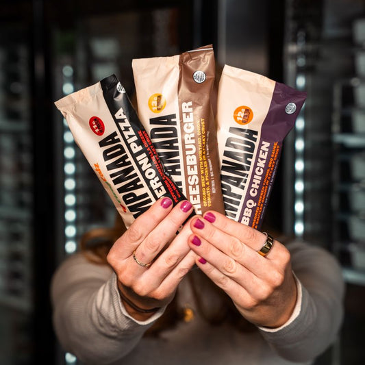 Person holding three Clean Eatz Empanada packages toward the camera—Pepperoni Pizza, Cheeseburger, and BBQ Chicken—with freezers in the background.