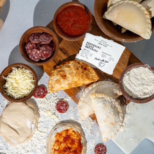 Top-down flat lay of Pepperoni Pizza empanada ingredients and prep: dough, flour, shredded cheese, sliced pepperoni, marinara sauce, and baked empanadas on a work surface.