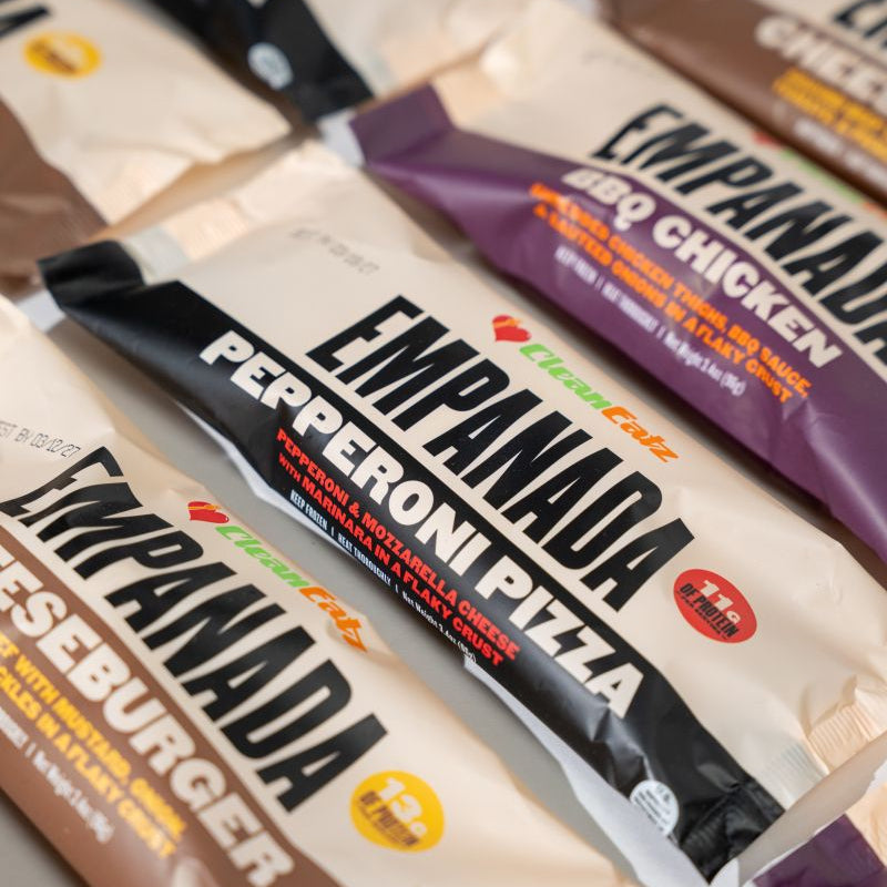 Close-up of assorted Clean Eatz Empanada packages laid out together, including Pepperoni Pizza, BBQ Chicken, and Cheeseburger flavors.