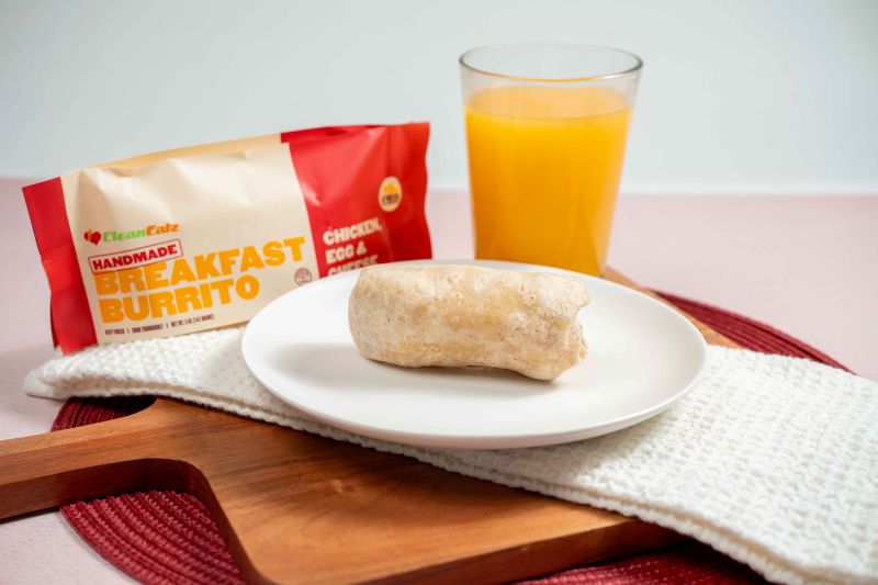 A plated breakfast burrito on a white plate with a glass of orange juice and the Chicken, Egg & Cheese burrito package in the background.