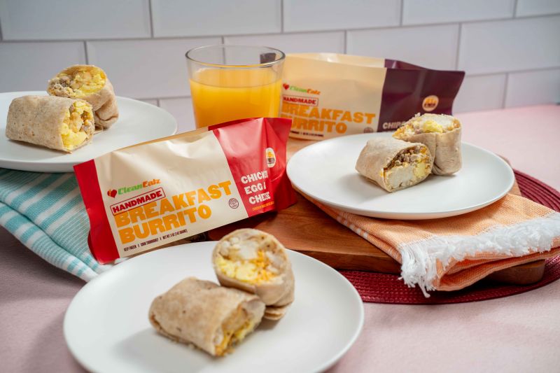 Several breakfast burritos—some cut open—on white plates with orange juice and multiple Clean Eatz Kitchen breakfast burrito packages on a tabletop.