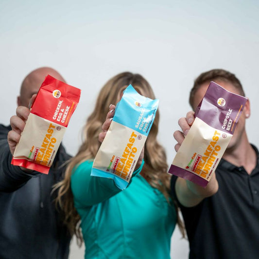 Three people holding up Clean Eatz Kitchen breakfast burrito packages—Chicken, Egg & Cheese, Sausage, Egg & Cheese, and Chipotle Beef—toward the camera.