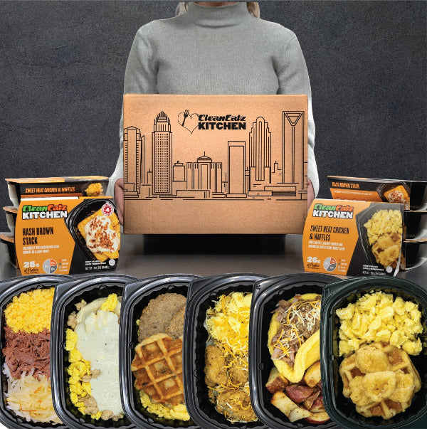 Clean Eatz Kitchen Breakfast Meal Plan — person holding branded shipping box with a lineup of breakfast entrées (e.g., hash brown stack, chicken & waffles, egg dishes) in trays and retail sleeves.