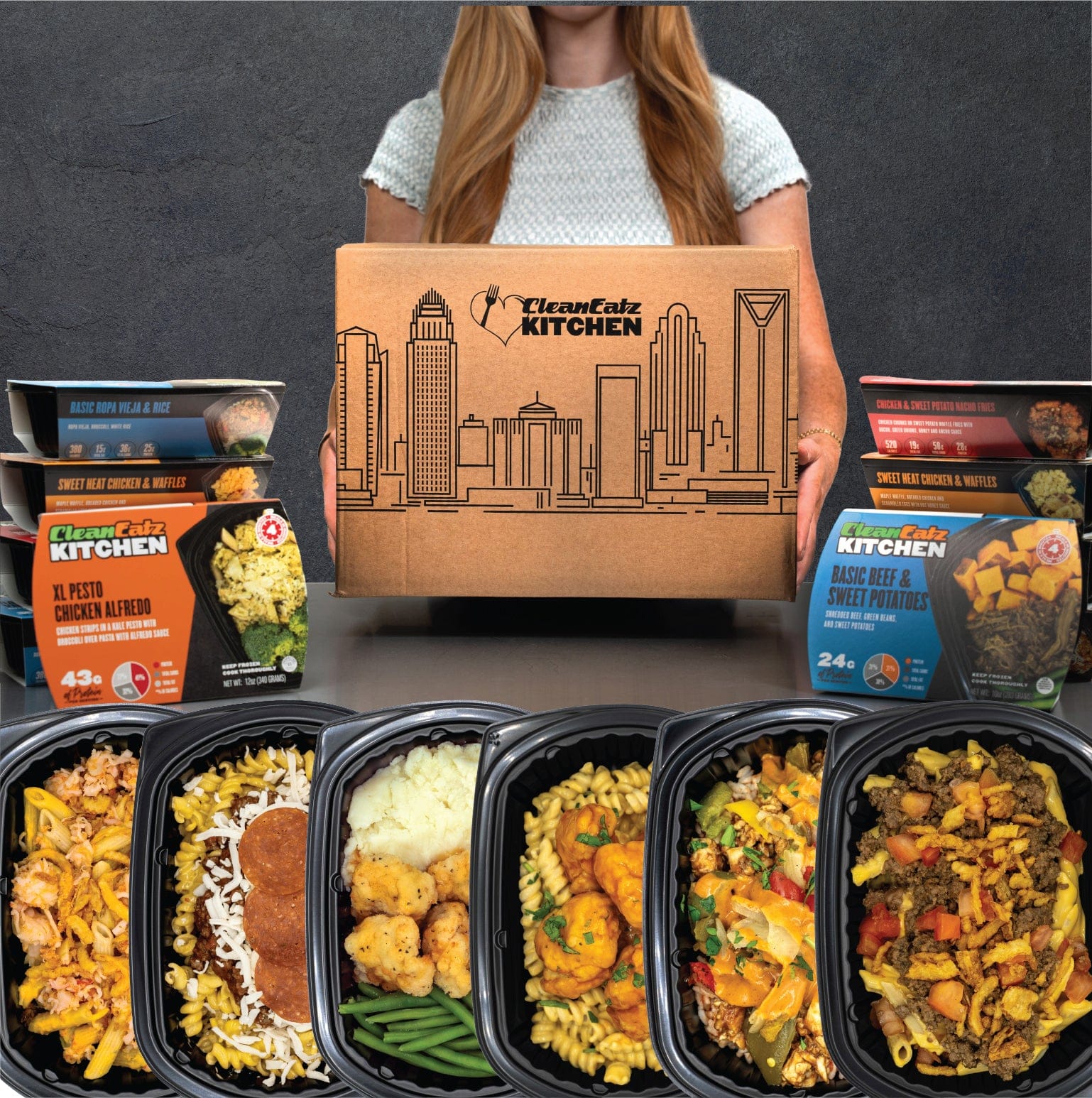 Clean Eatz Kitchen Build Your Meal Plan — person holding branded box with an assortment of prepared meals in trays and colorful entrée sleeves displayed in front.